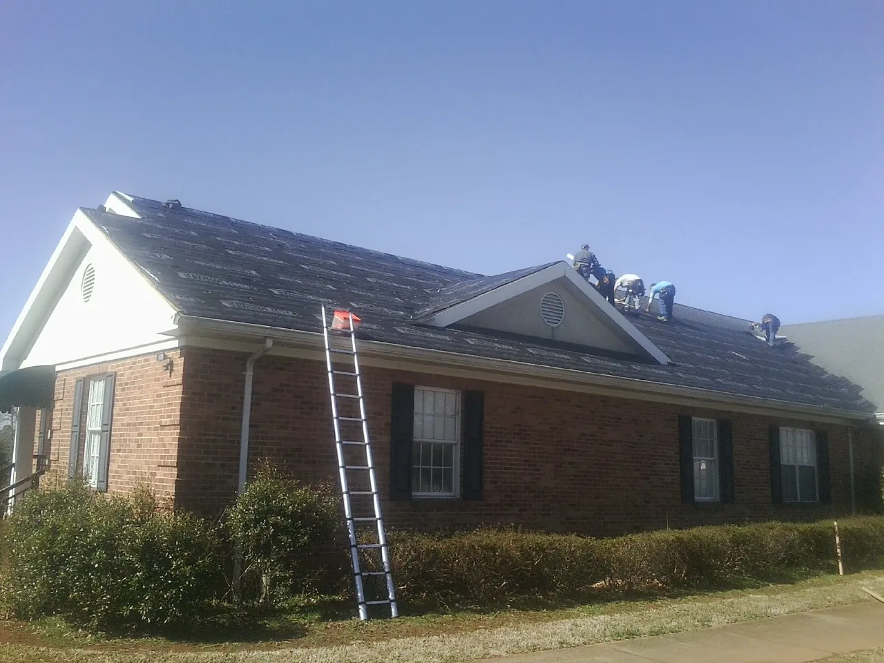 Crew tearing shingles off a brick ranch roof with a ladder set — Metal Roof Repair work by our crew in Slate Hill