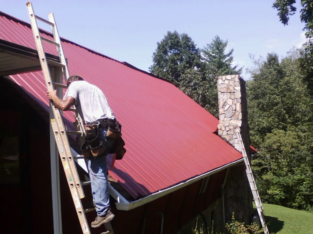 Roofer on a ladder beside a red metal roof and stone chimney — Soffit & Fascia Repair by Roofing Brothers in Slate Hill